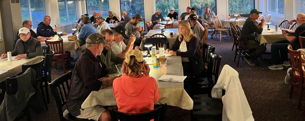 A gathering of people seated at tables in a dining area, engaged in conversations and activities, with natural light coming through large windows.