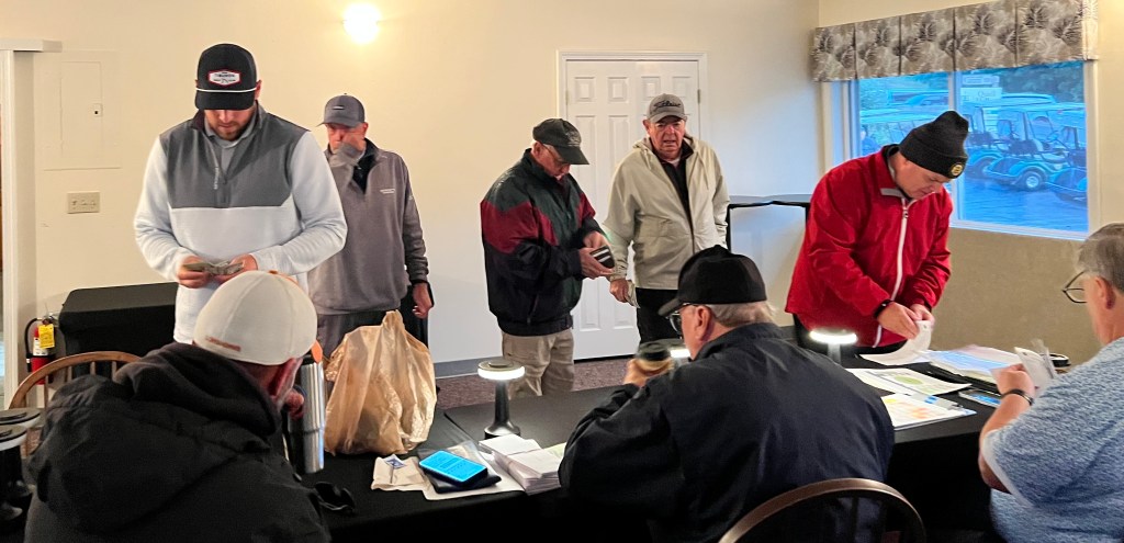 A group of men interacting at a registration table inside a building, with one man looking at his phone and others handling paperwork and cash.