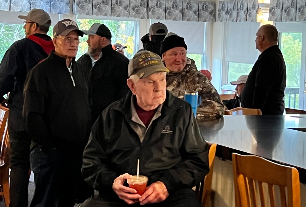 A gathering of several people indoors, with an elderly man sitting in the foreground holding a drink, while others are engaged in conversation in the background.