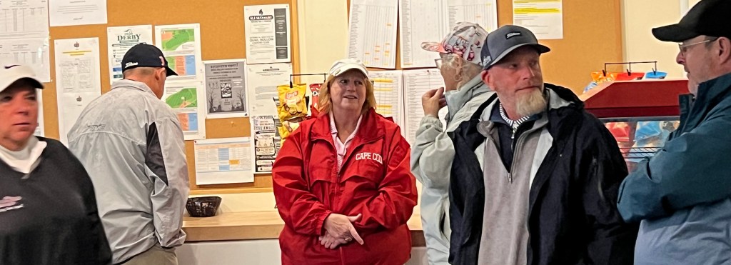 A group of people indoors at a counter, some interacting with a service area, with several notices and flyers pinned on a board behind them.