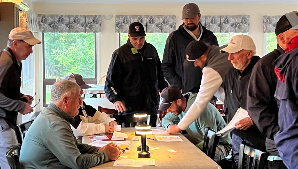 A group of men gathered around a table indoors, engaged in discussions and reviewing documents, with a lamp providing light. Windows in the background show greenery outside.