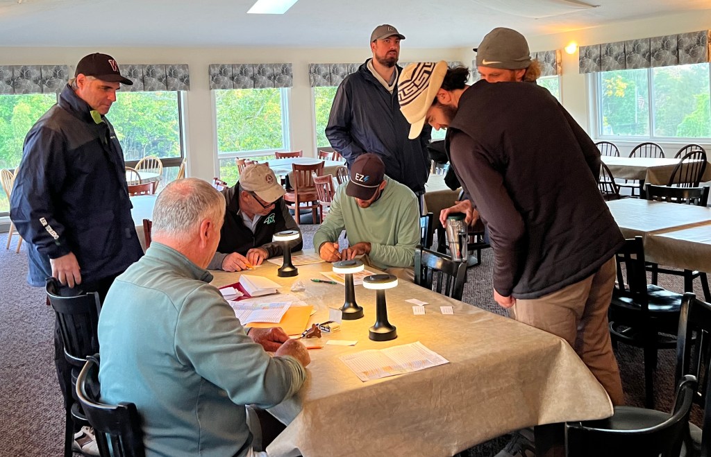 A group of six men interacting around tables in a well-lit dining area, with some working on paperwork and others observing, set against large windows with outside views.
