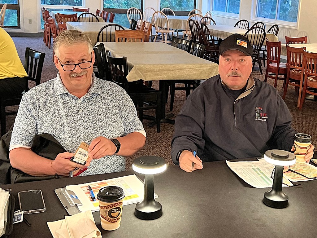 Two men sitting at a table in a well-lit room with wooden chairs and tables in the background. One man has glasses and is holding a box, while the other is holding a coffee cup and writing with a pen. Both are looking at the camera.